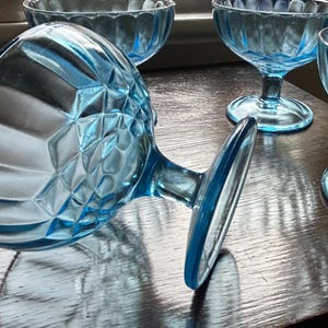 May include: A collection of vintage, light blue glass dessert dishes. The dishes have a diamond pattern on the bowl and a stem. The stem is attached to a round base. The dishes are arranged on a wooden surface.