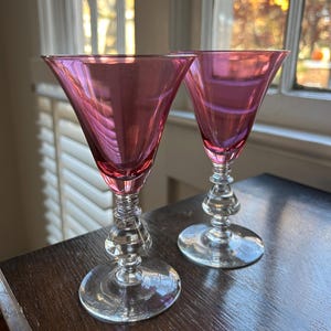 May include: Two vintage wine glasses with a pink hue. The glasses have a flared bowl design and a decorative stem with a clear, round base. The glasses are sitting on a dark wooden surface.