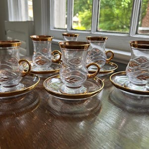 May include: Six clear glass teacups with gold rims and handles, each resting on a matching saucer. The cups have a decorative leaf design. The set is arranged on a dark wooden surface, with natural light from a window in the background.