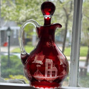 May include: A ruby red glass pitcher with a clear handle and stopper. The pitcher features etched white designs of trees and a building. The pitcher is sitting in front of a window.