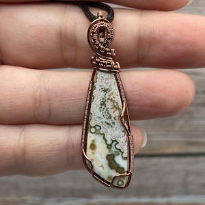 May include: A teardrop-shaped ocean jasper pendant featuring a complex, multi-coloured pattern of cream, olive green, and brown spots. The stone is encased in a decorative copper wire wrap with a spiral bail, suspended from a black cord necklace.