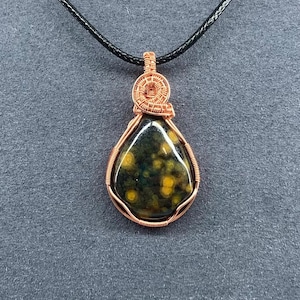 May include: A teardrop-shaped pendant with a dark green and yellow speckled stone, wrapped in copper wire. The wire is intricately woven at the top, and the pendant hangs from a black cord. The stone has a polished, glossy finish.