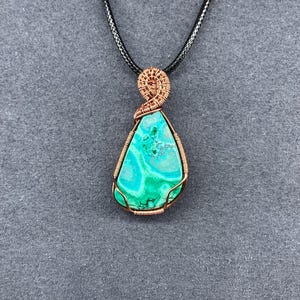 May include: A handcrafted pendant necklace featuring a teardrop-shaped, turquoise and green stone. The stone is wrapped in copper wire and suspended from a black cord. The wirework includes a decorative bail at the top.
