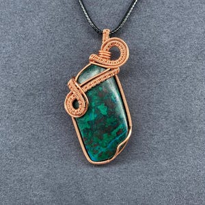 May include: A handcrafted pendant necklace featuring a rectangular, teal and green stone wrapped in copper wire. The pendant hangs from a black cord. The wire is intricately woven around the stone, creating a unique, artistic design.