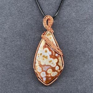May include: A teardrop-shaped pendant with a patterned stone, wrapped in copper wire, hangs from a black cord. The stone features a mix of brown, white, and yellow hues. The wirework forms a decorative frame and bail.
