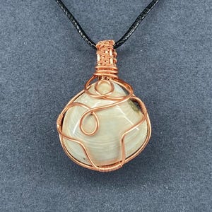 May include: A handcrafted pendant featuring a cream-colored, oval-shaped stone wrapped in copper wire. The wire is intricately woven around the stone, creating a decorative cage. The pendant hangs from a black cord, suitable for wearing.