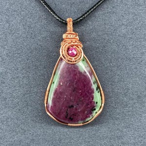 May include: A handcrafted pendant featuring a teardrop-shaped stone with a mix of deep red and green hues. The stone is wrapped in copper wire and accented with a small pink gemstone. The pendant hangs from a black cord.