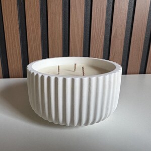 May include: A large, white, round candle in a ribbed concrete vessel. The candle has three wicks and a light-colored wax. The background features a wooden slat wall. This decorative candle is suitable for home decor.