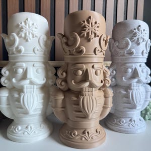 Puede incluir: Tres figuras decorativas de cascanueces en blanco, beige y gris. Cada una tiene una cara detallada, un sombrero alto con un copo de nieve y un uniforme tradicional. Las figuras están colocadas sobre una superficie blanca, con una casa de jengibre y decoración navideña en el fondo.