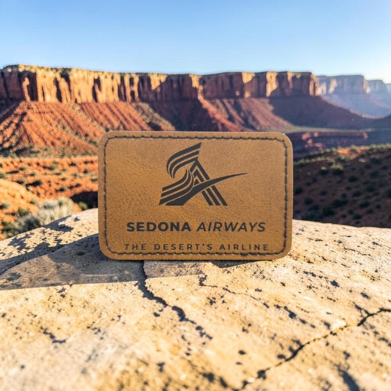 May include: A rectangular tan leather patch with rounded corners and stitched edges. The patch features a black logo of stylized wings and the text "SEDONA AIRWAYS" followed by "THE DESERT'S AIRLINE" printed in a clean, sans-serif font.