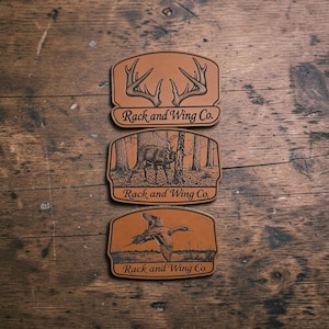 May include: Three tan leather patches with black engraved designs. The top patch shows deer antlers, the middle shows a deer in a forest, and the bottom shows a flying duck. Each patch features the text Rack and Wing Co. in black script.