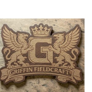 May include: A brown and tan wooden emblem featuring two winged griffins flanking a large letter "G" topped with a crown. The text "GRIFFIN FIELDCRAFT" is displayed on a banner below the griffins.