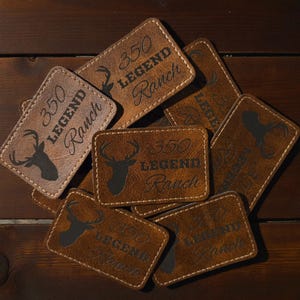 May include: A collection of rectangular brown patches with stitched edges. Each patch features a dark silhouette of a deer head and the text 350 LEGEND Ranch engraved on the surface. The patches are arranged in a overlapping pile.