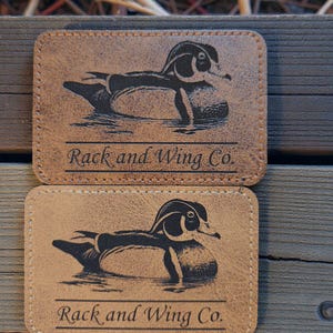 May include: Two rectangular tan leather patches with rounded corners, each featuring a black laser-engraved illustration of a wood duck swimming and the text "Rack and Wing Co." printed in a serif font above a horizontal line.
