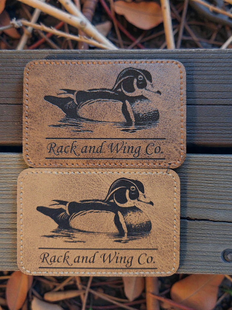May include: Two rectangular tan leather patches with rounded corners, each featuring a black laser-engraved illustration of a wood duck swimming and the text "Rack and Wing Co." printed in a serif font above a horizontal line.