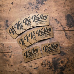 May include: A collection of rectangular tan patches featuring the black printed text Valley Speedway Grain Valley, MO. Each patch has rounded corners and a textured surface, arranged in a scattered overlapping pattern on a wooden surface.