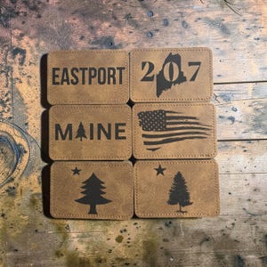 May include: Six rectangular tan faux leather patches with dark brown laser-engraved designs. The patches feature the text EASTPORT, the number 207 with a Maine state outline, the text MAINE with a tree, a distressed American flag, and two pine trees.