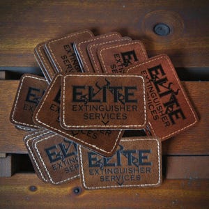 May include: A collection of rectangular brown leather patches featuring white stitched borders. Each patch is embossed with the black text ELITE EXTINGUISHER SERVICES and a graphic icon of a flame above the word ELITE.