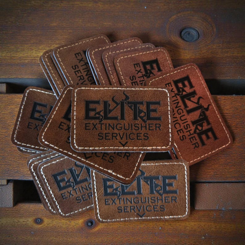 May include: A collection of rectangular brown leather patches featuring white stitched borders. Each patch is embossed with the black text ELITE EXTINGUISHER SERVICES and a graphic icon of a flame above the word ELITE.