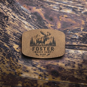 May include: A tan rectangular patch with rounded corners featuring a black engraved graphic of a moose standing in front of a forest and mountains. The text reads FOSTER MOOSE CAMP 2026 with decorative flourishes above and below the year.
