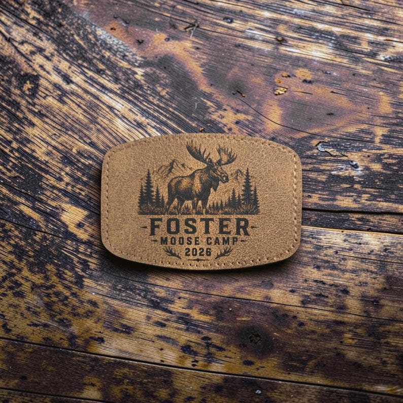 May include: A tan rectangular patch with rounded corners featuring a black engraved graphic of a moose standing in front of a forest and mountains. The text reads FOSTER MOOSE CAMP 2026 with decorative flourishes above and below the year.