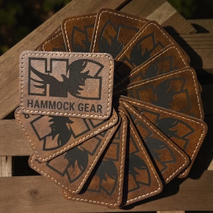 May include: A collection of brown leather patches with the text "HAMMOCK GEAR" and a stylized bird logo. The patches are arranged in a circular pattern, showcasing their design and texture. The patches have a stitched border.