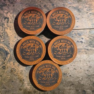 May include: Five circular tan leather patches with stitched edges, each featuring a dark brown embossed graphic of hikers in a mountain landscape. The text reads WALK WITH ME, YOUNG MEN'S CAMP, and 2026, arranged in a circular design.