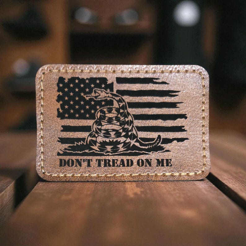 May include: A rectangular tan leather patch with rounded corners and visible stitching along the edges. The surface features a laser-engraved black design of a coiled rattlesnake over a distressed American flag with the text DON'T TREAD ON ME.