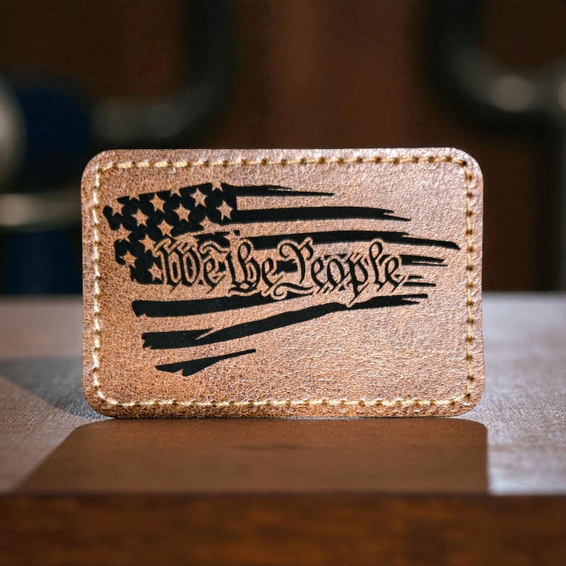 May include: A rectangular tan leather patch with rounded corners and light-colored perimeter stitching. The surface features a black laser-engraved design of a distressed American flag with the text We the People centered across the stripes.