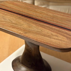 Könnte beinhalten: Ein handgefertigter Beistelltisch aus Holz mit rechteckiger Platte und Säulenfuß. Die Tischplatte hat eine dunkle Streifeneinlage. Der Tisch ist aus warmtonigem Holz gefertigt, das die natürliche Maserung zeigt. Der Tisch ist ca. 76 cm lang.