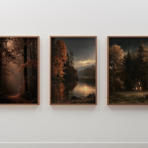 May include: A set of three framed wall art prints featuring autumn forest landscapes. The left print shows a sunlit path, the center depicts a lake at sunset, and the right displays a small cabin nestled in a dark, misty, golden-toned forest.