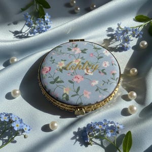 Peut inclure: Un miroir de poche rond avec un motif floral et le nom "Ashley" en lettres dorées. Le miroir est posé sur un fond de satin bleu clair, orné de perles et de fleurs myosotis. Le compact a un cadre doré.
