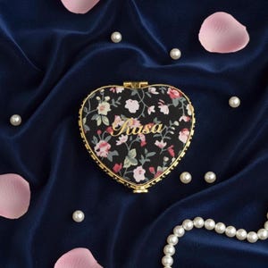 May include: Heart-shaped compact mirror with a floral pattern on a black background. The mirror has a gold-colored frame and the word "Rasa" in gold script. The background is a dark blue fabric with scattered pearls and pink petals.