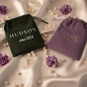 May include: Two small drawstring bags, one dark green and one purple, rest on a satin surface with scattered pearls and purple flowers. The green bag has the text "HUDSON since 2024" in white. The purple bag has the text "BRAND COLOR" in gold.
