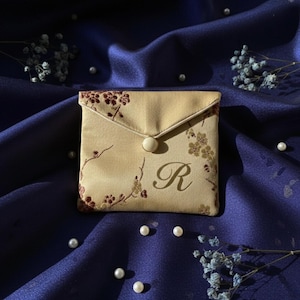 May include: A small, beige fabric pouch with a floral pattern and the letter "R" printed on the front. The pouch has a flap closure secured by a white button. The background is a dark blue fabric with scattered pearls and small blue flowers.