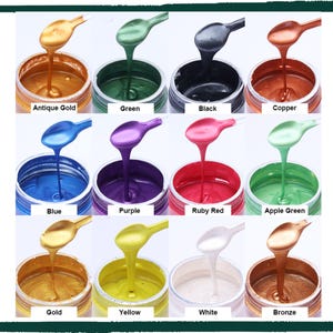 May include: A grid of twelve small jars containing metallic paint in various colors, each with a matching spoon dripping paint. Colors include antique gold, green, black, copper, blue, purple, ruby red, apple green, gold, yellow, white, and bronze.