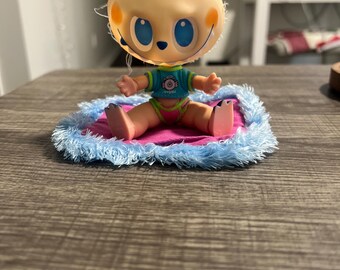 May include: A small, tan-coloured doll with large blue eyes and a smiling expression. The doll is wearing a teal shirt and pink underwear, sitting on a pink and blue fluffy mat. The doll's arms are outstretched.