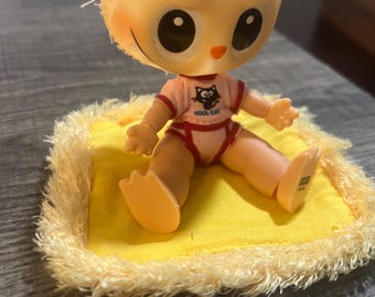 May include: A vintage doll with a large, round head and big eyes, wearing a pink onesie with a cat graphic. The doll is sitting on a yellow mat with a fluffy, fringed border. The text "KOOL KAT" is visible on the onesie.