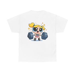May include: White t-shirt featuring a cartoon character with blonde pigtails lifting a barbell. The character is wearing pink shorts and black shoes. The barbell has blue weights. The design includes sparkling star accents.