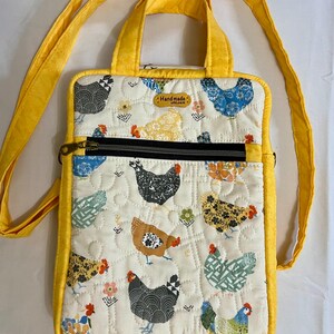 May include: A quilted crossbody bag with a yellow strap and trim. The bag features a cream-colored fabric with a repeating pattern of colorful chickens and flowers. A black zipper pocket is visible on the front, and the bag has a small handle. The tag reads "Handmade".