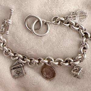 Puede incluir: Pulsera de plata con dijes que incluye un s&iacute;mbolo de d&oacute;lar, dados, naipes y una moneda. La pulsera tiene un cierre de palanca y se muestra sobre una superficie suave y texturizada.