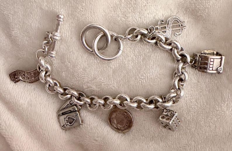 Puede incluir: Pulsera de plata con dijes que incluye un s&iacute;mbolo de d&oacute;lar, dados, naipes y una moneda. La pulsera tiene un cierre de palanca y se muestra sobre una superficie suave y texturizada.