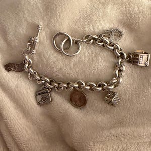 Puede incluir: Una pulsera de plata con dijes variados, incluyendo un signo de d&oacute;lar, dados, naipes y una moneda. La pulsera tiene un cierre de palanca y un dise&ntilde;o de cadena gruesa. Los dijes son detallados.