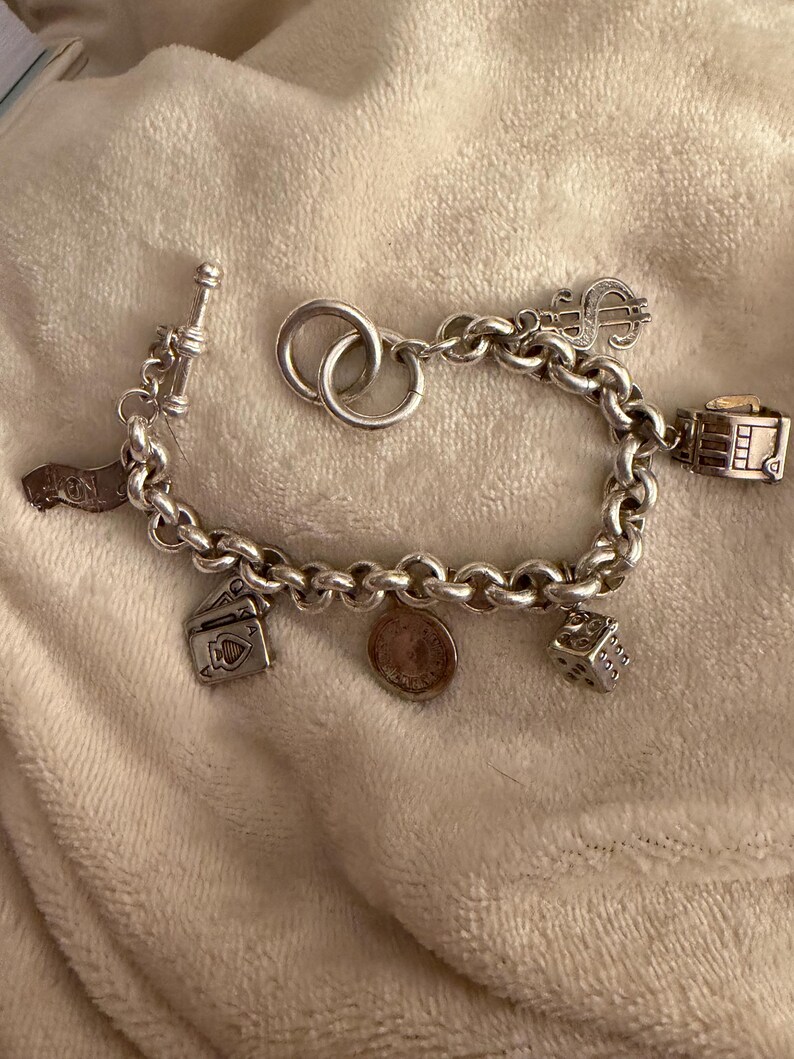 Puede incluir: Una pulsera de plata con dijes variados, incluyendo un signo de d&oacute;lar, dados, naipes y una moneda. La pulsera tiene un cierre de palanca y un dise&ntilde;o de cadena gruesa. Los dijes son detallados.