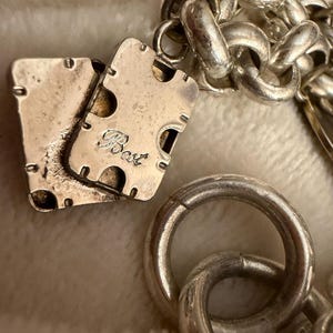 Puede incluir: Primer plano de un collar plateado con una cadena gruesa y un cierre rectangular. El cierre tiene la palabra "Best" grabada. El collar est&aacute; sobre una superficie blanca y suave.