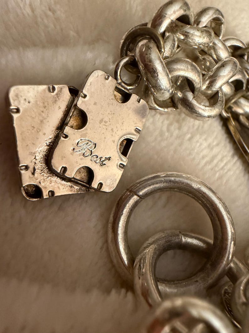 Puede incluir: Primer plano de un collar plateado con una cadena gruesa y un cierre rectangular. El cierre tiene la palabra "Best" grabada. El collar est&aacute; sobre una superficie blanca y suave.