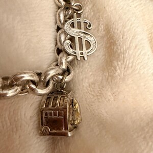 Puede incluir: Pulsera de plata con un dije de signo de d&oacute;lar y una m&aacute;quina tragamonedas. La pulsera tiene un dise&ntilde;o de cadena gruesa. El dije del signo de d&oacute;lar es detallado, y el dije de la m&aacute;quina tragamonedas tiene un interior dorado.