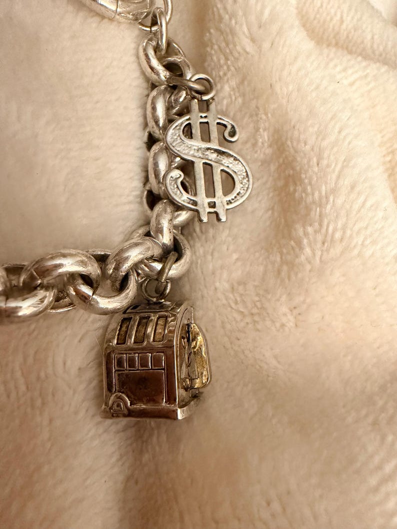 Puede incluir: Pulsera de plata con un dije de signo de d&oacute;lar y una m&aacute;quina tragamonedas. La pulsera tiene un dise&ntilde;o de cadena gruesa. El dije del signo de d&oacute;lar es detallado, y el dije de la m&aacute;quina tragamonedas tiene un interior dorado.