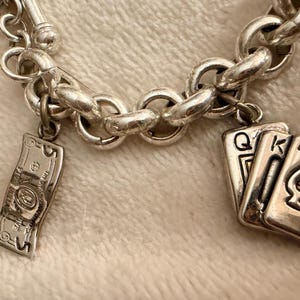 Puede incluir: Pulsera de abalorios plateada con una serie de eslabones redondos. La pulsera presenta tres colgantes: un billete de banco en miniatura detallado y dos naipes, la Reina y el As de Picas.
