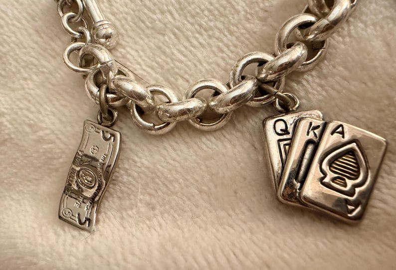 Puede incluir: Pulsera de abalorios plateada con una serie de eslabones redondos. La pulsera presenta tres colgantes: un billete de banco en miniatura detallado y dos naipes, la Reina y el As de Picas.